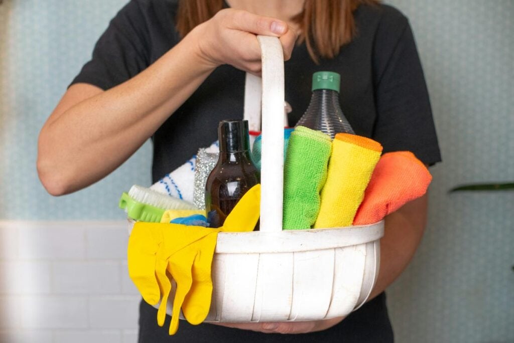 A woman holding a portable cleaning caddy filled with colorful microfiber cloths, cleaning brushes, spray bottles, and yellow rubber gloves, ideal for efficient cleaning.