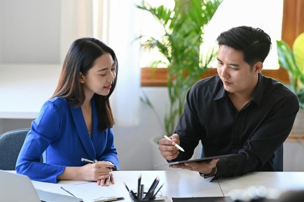 A woman and a man sitting at a desk in a bright, modern office. The man is showing the woman information on a digital tablet, while she takes notes. Both look focused and engaged, suggesting a financial planning or budgeting discussion.