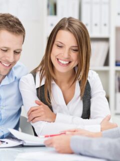 A smiling couple sitting at a table across from a professional advisor. The advisor is showing them paperwork as they review documents together, highlighting a positive and collaborative financial planning meeting.