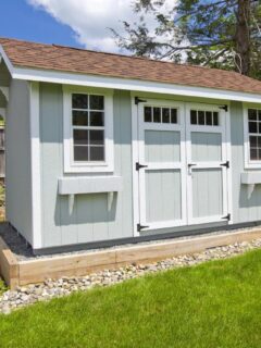 Light gray storage shed with white trim and double doors, surrounded by a landscaped yard and stone accents, ideal for outdoor storage shed organization.