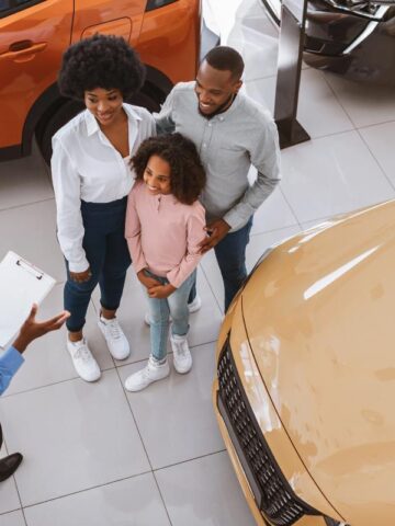 A smiling family with a young daughter talks to a car salesman in a dealership showroom. They are exploring SUV options similar to a used Dodge Durango for family driving.