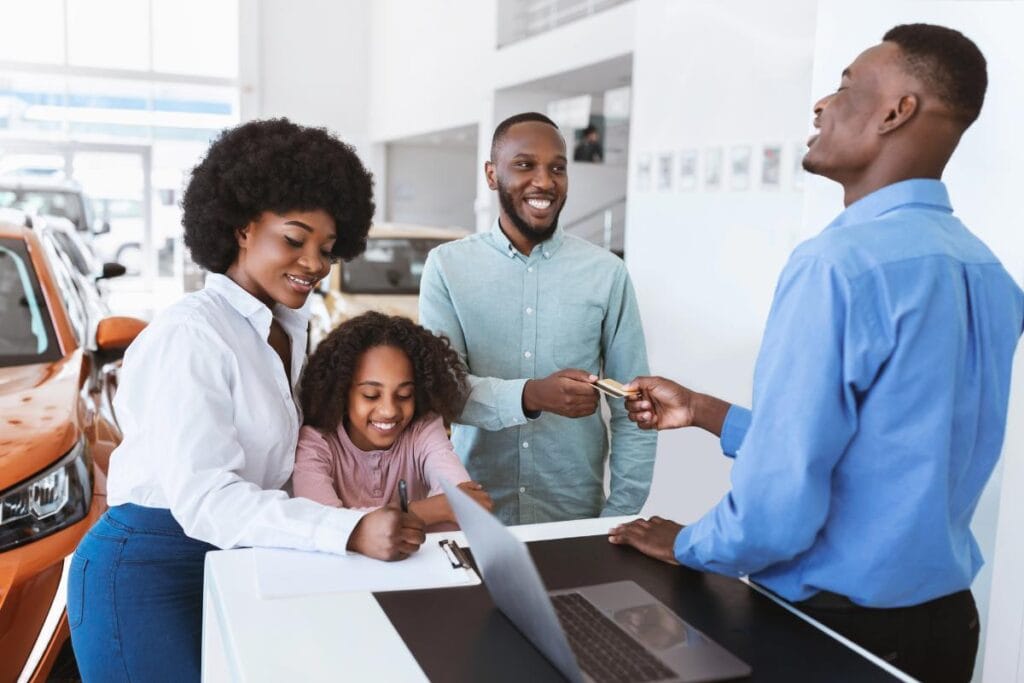 A cheerful family completes paperwork and makes a payment to a car salesman at a dealership. They are finalizing the purchase of a family-friendly SUV like a used Dodge Durango.
