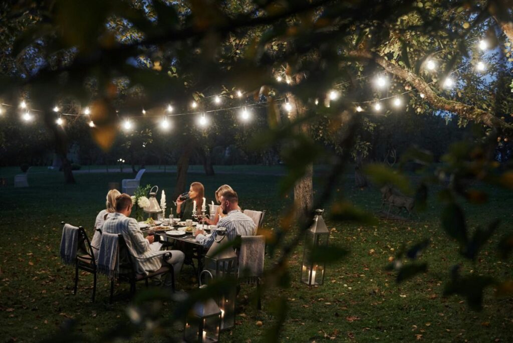 Friends enjoying an outdoor dinner at dusk beneath warm string lights in a leafy garden.