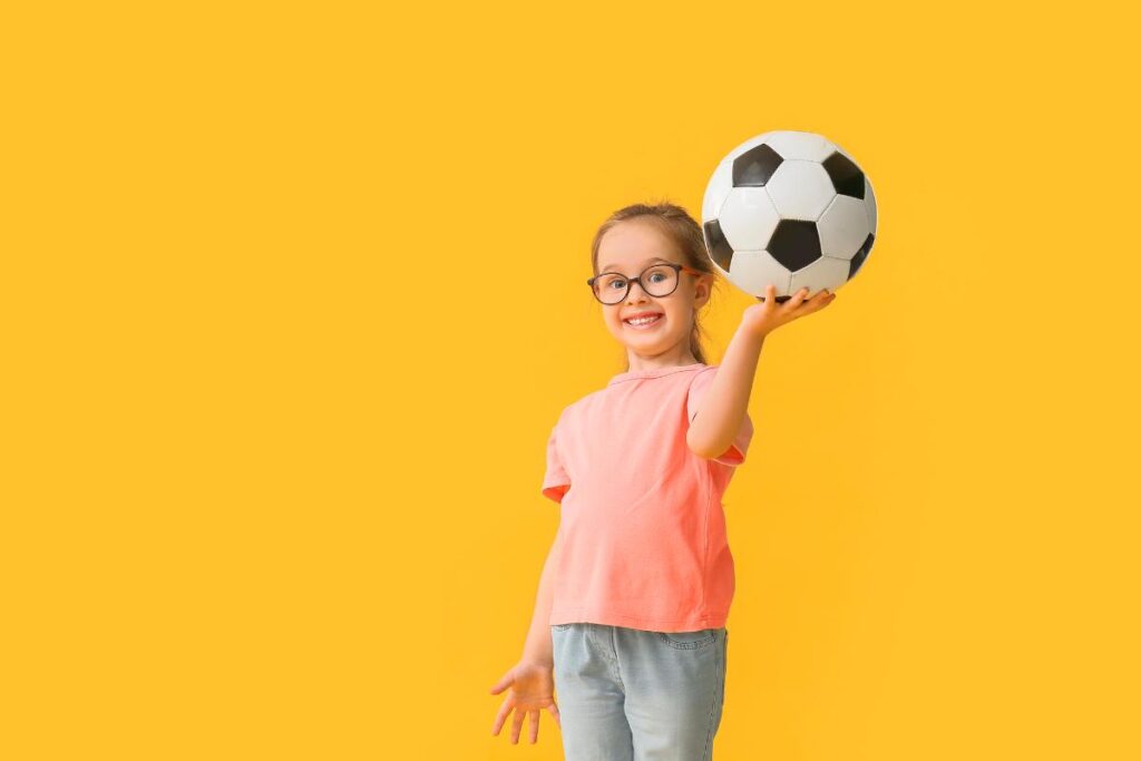 Smiling young girl wearing glasses and a pink shirt holding a soccer ball, standing in front of a bright yellow background.