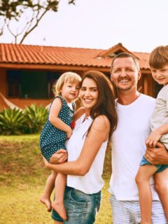 Smiling family of four standing outside their home on a sunny day, with parents holding their two young children in front of a terracotta-colored house with a tiled roof and green lawn.