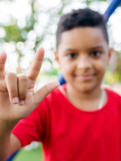 Smiling hearing-impaired boy in a red shirt making the “I love you” handshape in sign language at an inclusive outdoor playground, symbolising children learning effective sign-language communication.