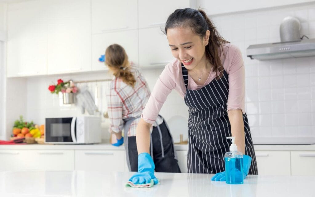 Two professional cleaners wearing aprons and gloves efficiently wiping down surfaces in a bright, modern kitchen, highlighting professional cleaning services.