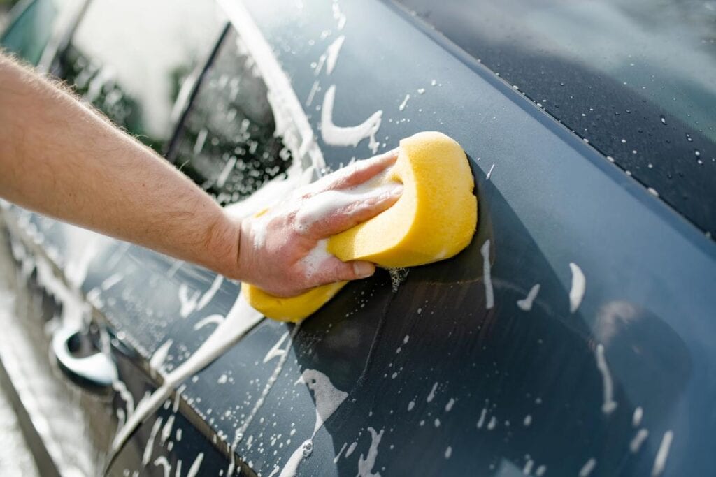 Close-up of a person washing a car door with a yellow sponge and soapy water, showing proper cleaning technique.
