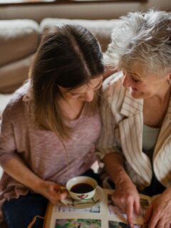 Senior mother and adult daughter sitting on a cozy couch, looking at a photo album and bonding in a multigenerational living space.