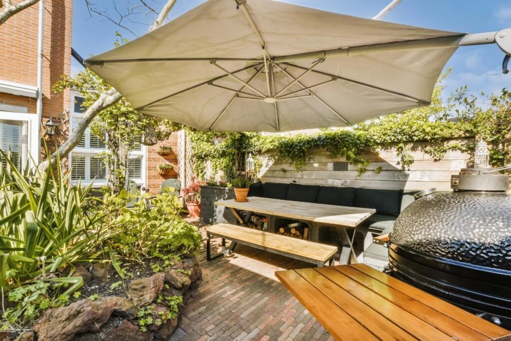 Sunny patio featuring a large shade umbrella over a timber dining set surrounded by greenery.