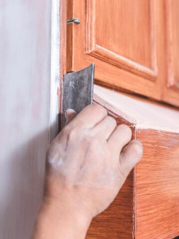Hand sanding wooden kitchen cabinets to prepare the surface for painting during a budget-friendly kitchen cabinet upgrade project.