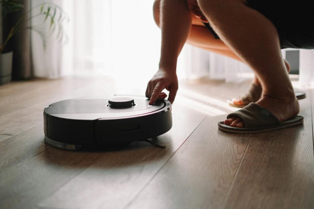 Person activating a robotic vacuum cleaner on a hardwood floor, highlighting smart technology and automated cleaning to save time in hectic lifestyles.