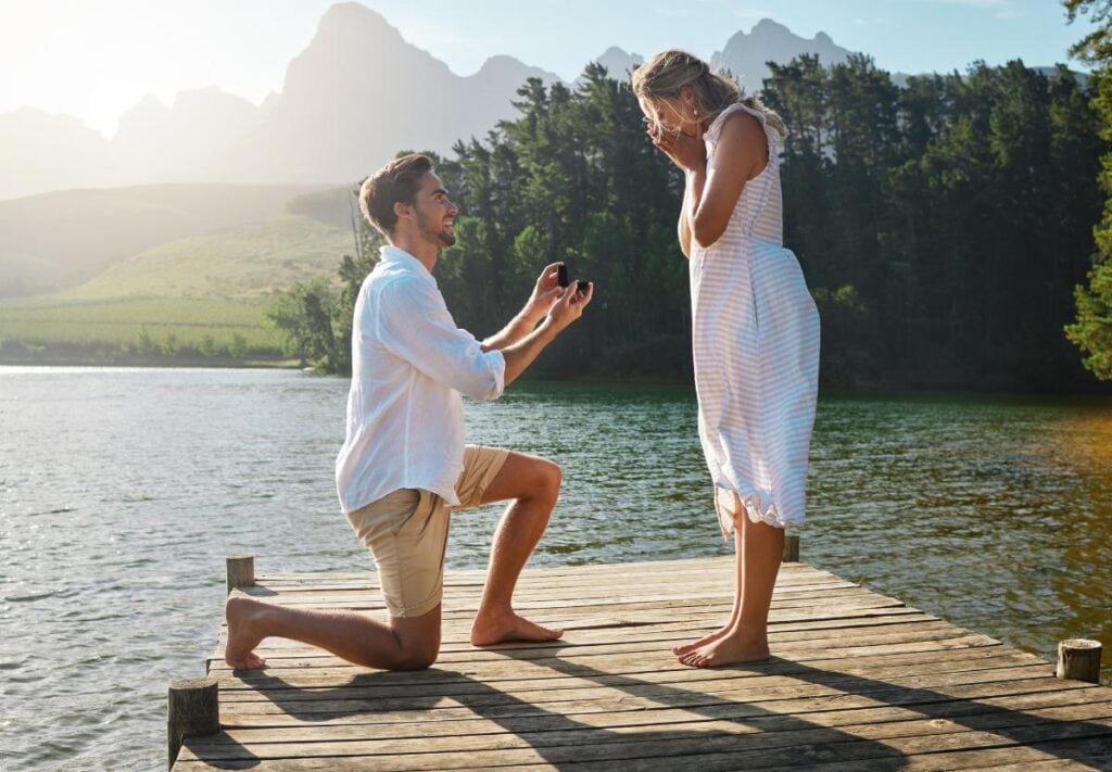 A man kneeling on a wooden dock by a scenic lake, proposing to a surprised woman in a white sundress, with forested hills and mountains in the background.