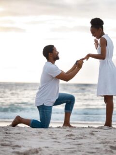 A man kneeling on the sand at the beach, holding a ring box and placing a ring on a woman’s finger as she smiles with excitement at sunset.
