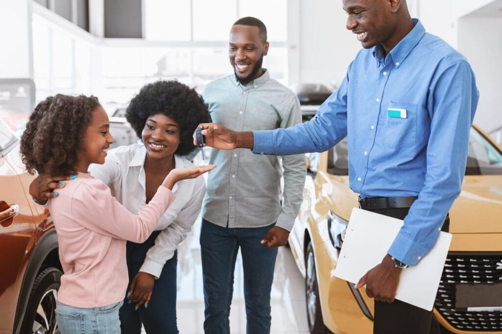 A car salesman hands over keys to a happy young girl standing with her family next to an SUV in a showroom, celebrating getting a versatile vehicle such as a used Dodge Durango.