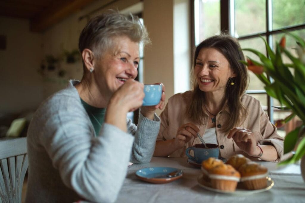 Smiling older woman and daughter drinking coffee and eating muffins at a kitchen table, showing connection and warmth in multigenerational households.