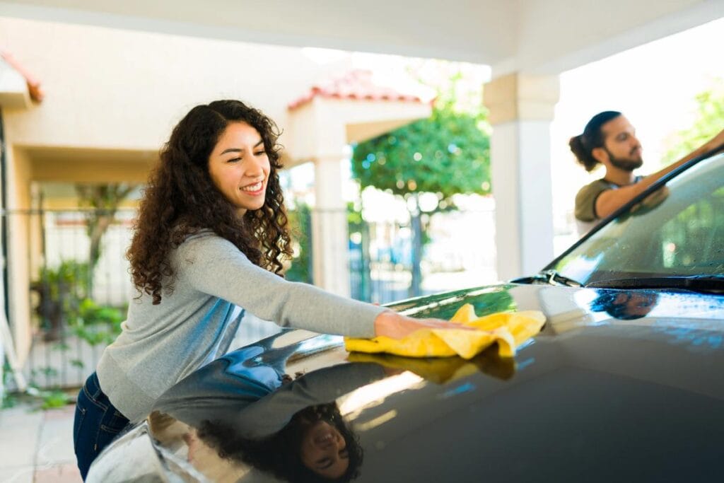 A cheerful woman drying a car hood with a yellow cloth while a man cleans the windshield in the background.