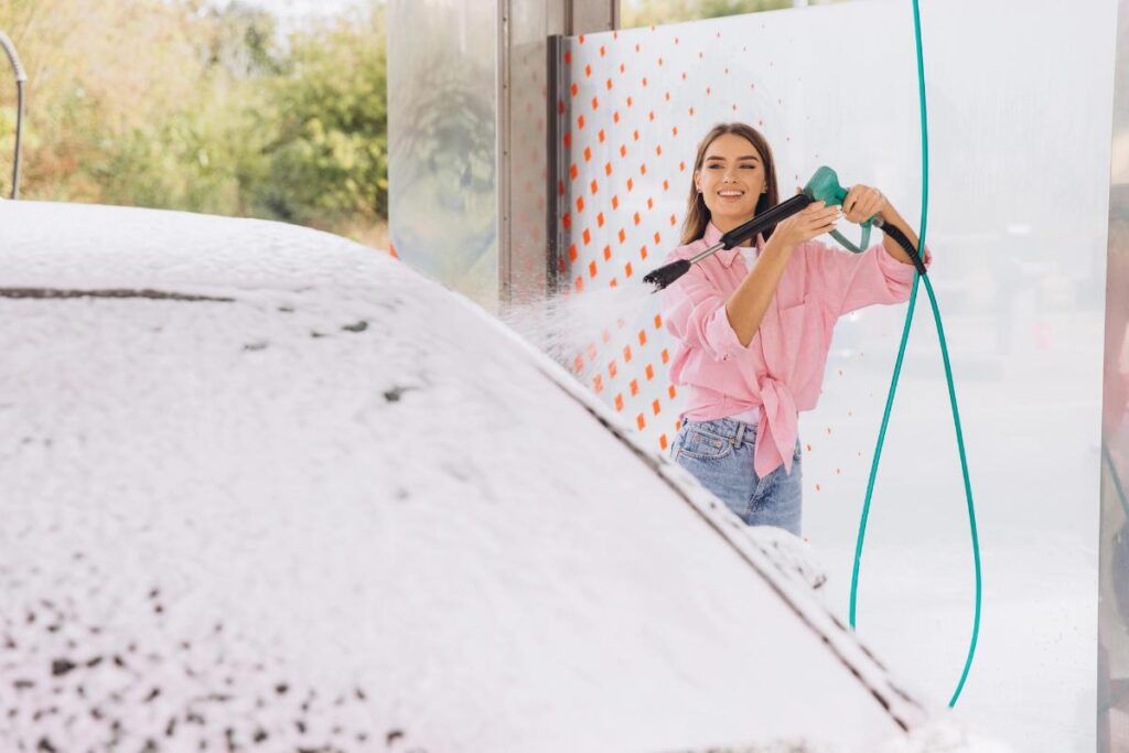 A young woman at a car wash using a pressure washer to rinse thick foam off her car, demonstrating the pre-wash rinse step.