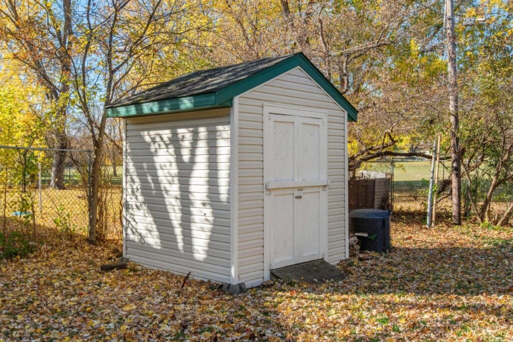 Beige backyard storage shed with green roof and white doors, located among autumn trees, perfect for showcasing storage shed organization ideas.