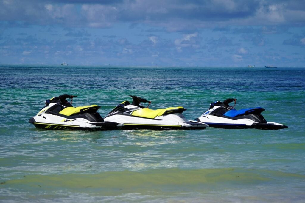 Three jet skis with bright yellow and blue seats floating on calm turquoise water under a partly cloudy sky.