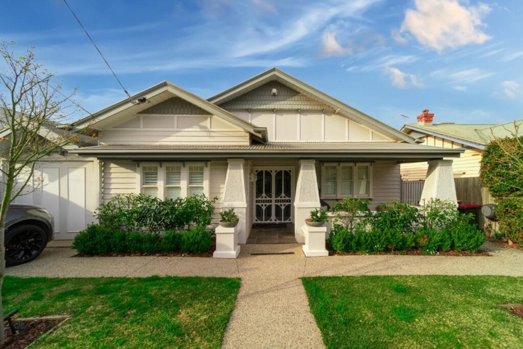 Front view of a charming white weatherboard house with a gabled roof, neatly trimmed hedges, and a landscaped front yard with a paved path leading to the entrance.