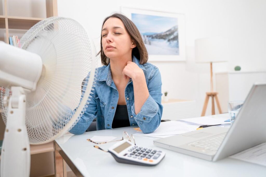 Young woman cooling off with an electric fan while working from home to reduce air conditioning costs and save energy.