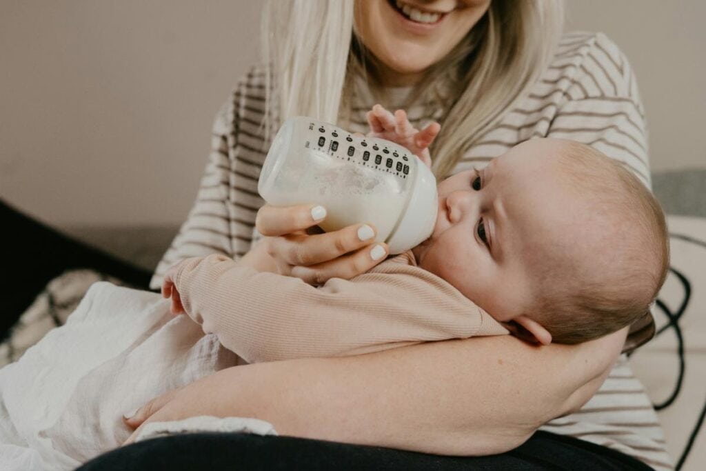 Smiling mother bottle feeding infant while holding baby in her arms; baby dressed in soft beige pajamas and cuddling a blanket.