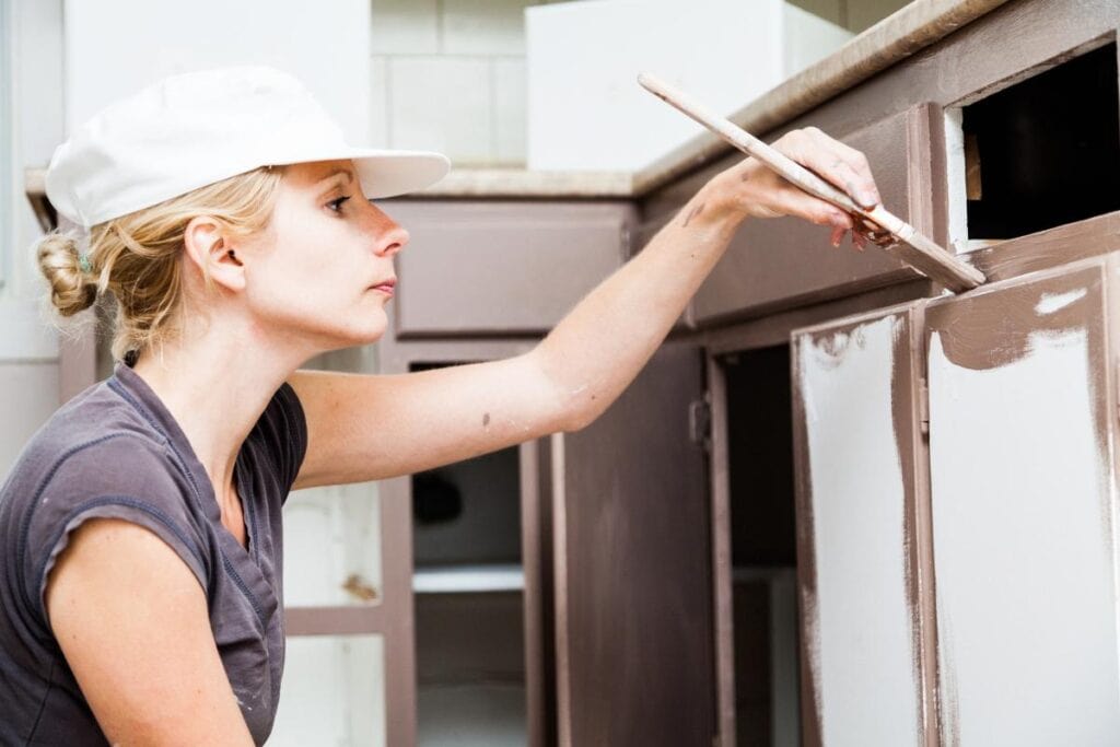 Woman painting brown kitchen cabinets with a brush as part of a DIY kitchen cabinet makeover to refresh the space.