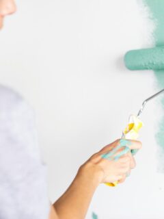 Person using a roller to apply teal paint on a white wall, demonstrating proper painting technique for a smooth, professional finish.