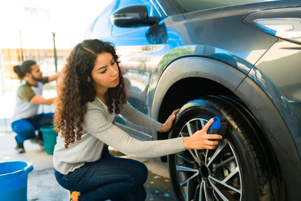 A woman cleaning a car wheel with a brush, focusing on tire care, while a man washes another part of the car in the background.