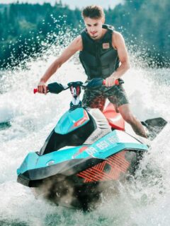 A young man wearing a life vest riding a blue and red jet ski at high speed, creating a large splash on a sunny day with forested hills in the background.