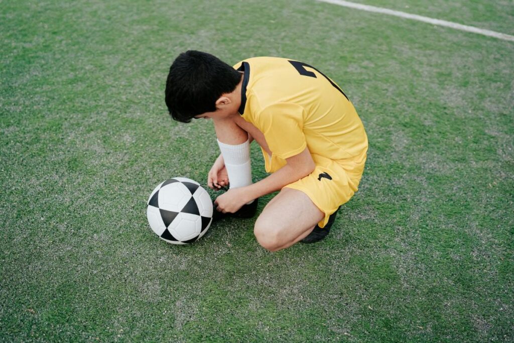 Child in a yellow soccer uniform and glasses kneeling on a soccer field, tying their shoe next to a black and white soccer ball.