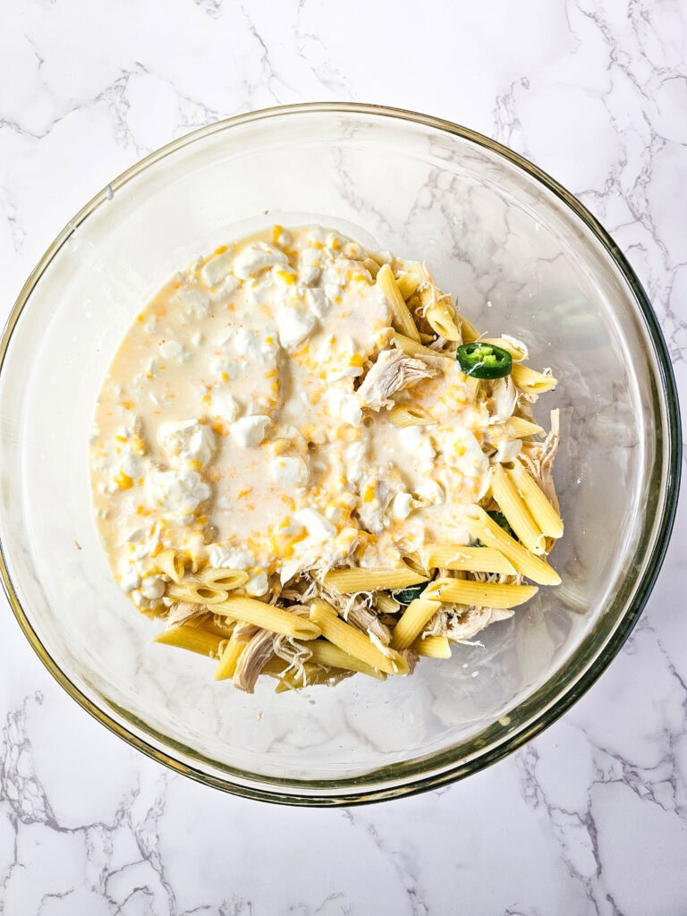 Cooked penne and shredded chicken in a large bowl with creamy cheese sauce and jalapeño slices, ready to stir for jalapeño popper chicken casserole.