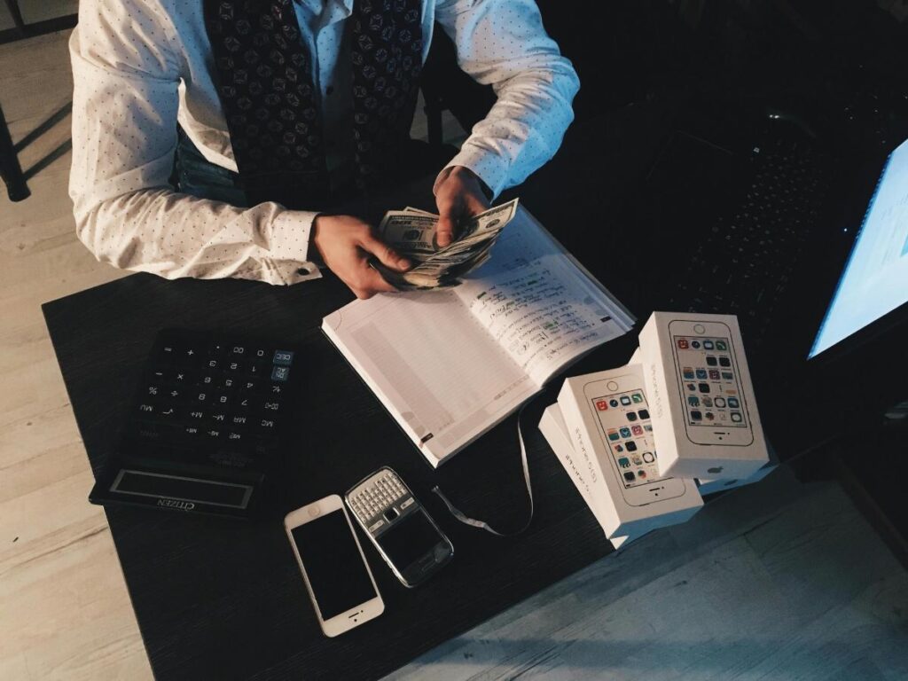 Entrepreneur managing small business finances by counting cash at a desk with a calculator, smartphone boxes, and a ledger notebook.