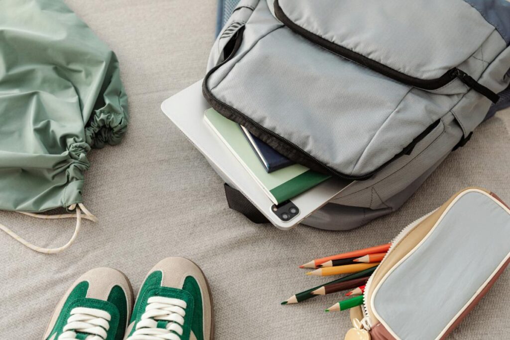 Close-up of a gray school backpack packed with notebooks, a tablet, and pencils, surrounded by sneakers and a drawstring bag—ready for back-to-school prep.