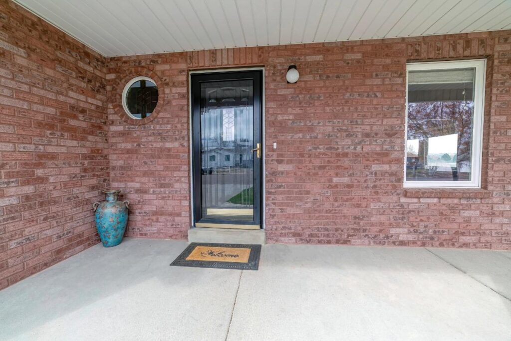 Front porch of a home with red brick exterior, black storm door, welcome mat, and ceramic vase, showing well-maintained brickwork and mortar joints.