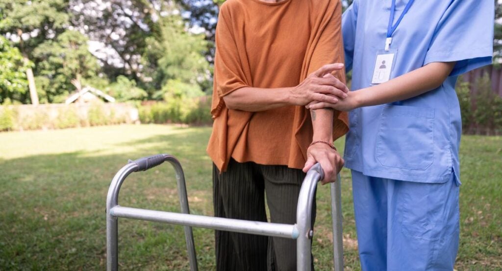 Support worker assisting an elderly woman with a walker outdoors, offering gentle arm support in a garden setting.