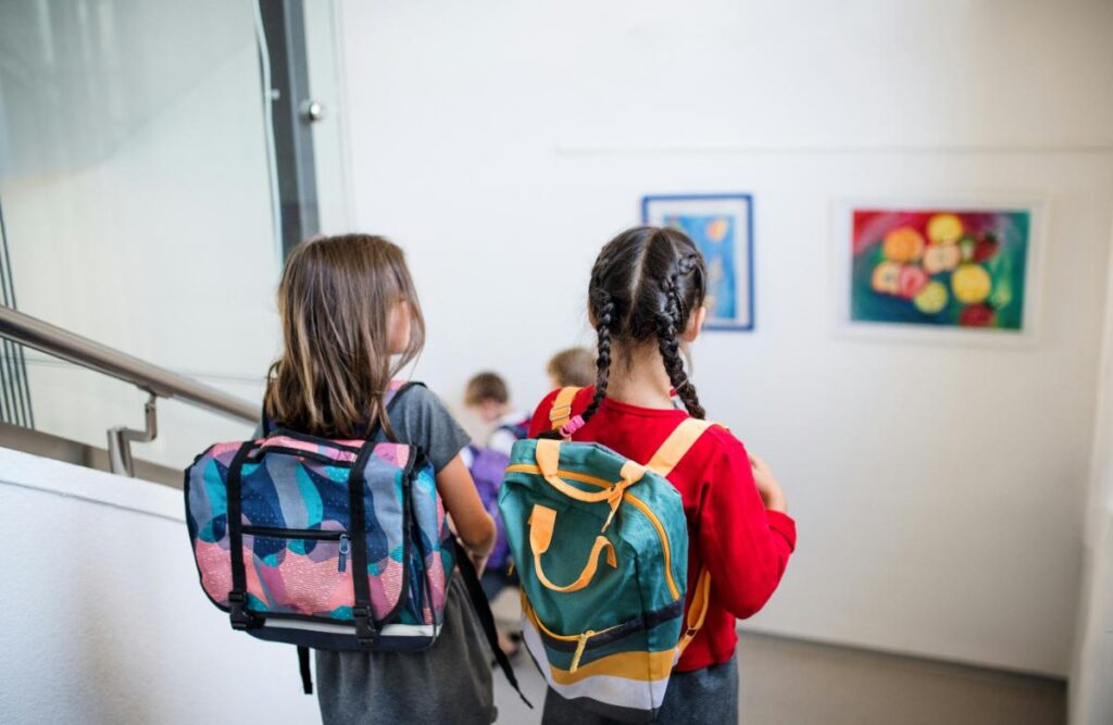 Two young schoolgirls with backpacks walking down a school hallway, representing back-to-school excitement and the start of a new school year.