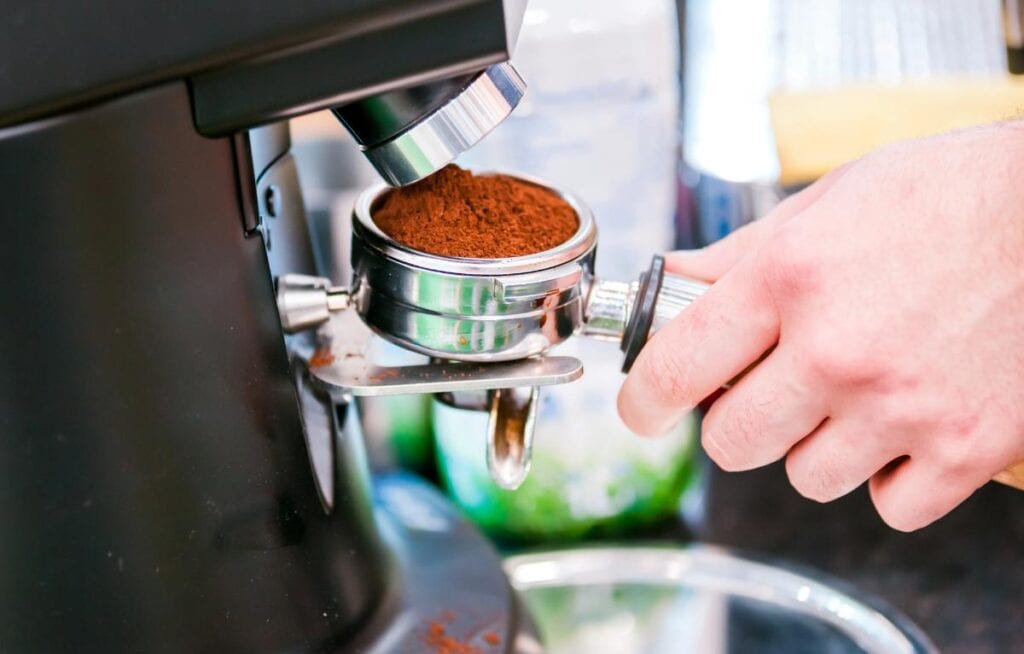 A hand holding a portafilter being filled with finely ground coffee beans, emphasizing freshness and grind quality for espresso preparation.