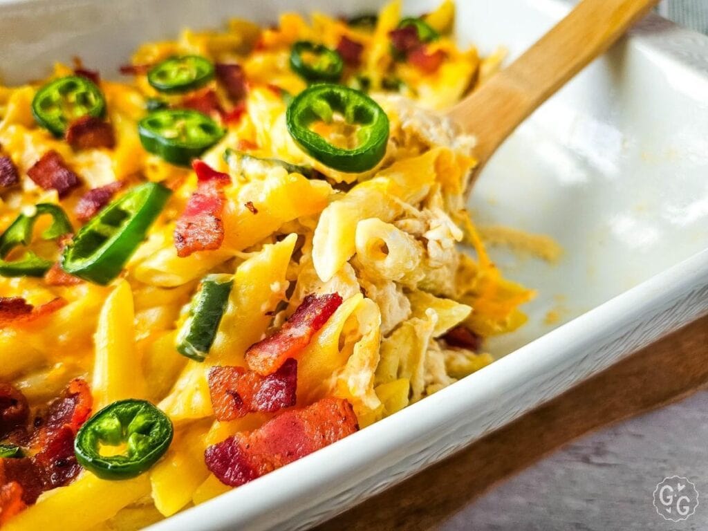 Cheesy jalapeño popper chicken pasta bake in the casserole dish with a wooden spoon and bacon pieces.