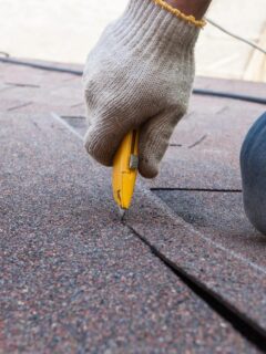 Close-up of a roofing contractor cutting asphalt shingles with a utility knife during roof repair and maintenance work.