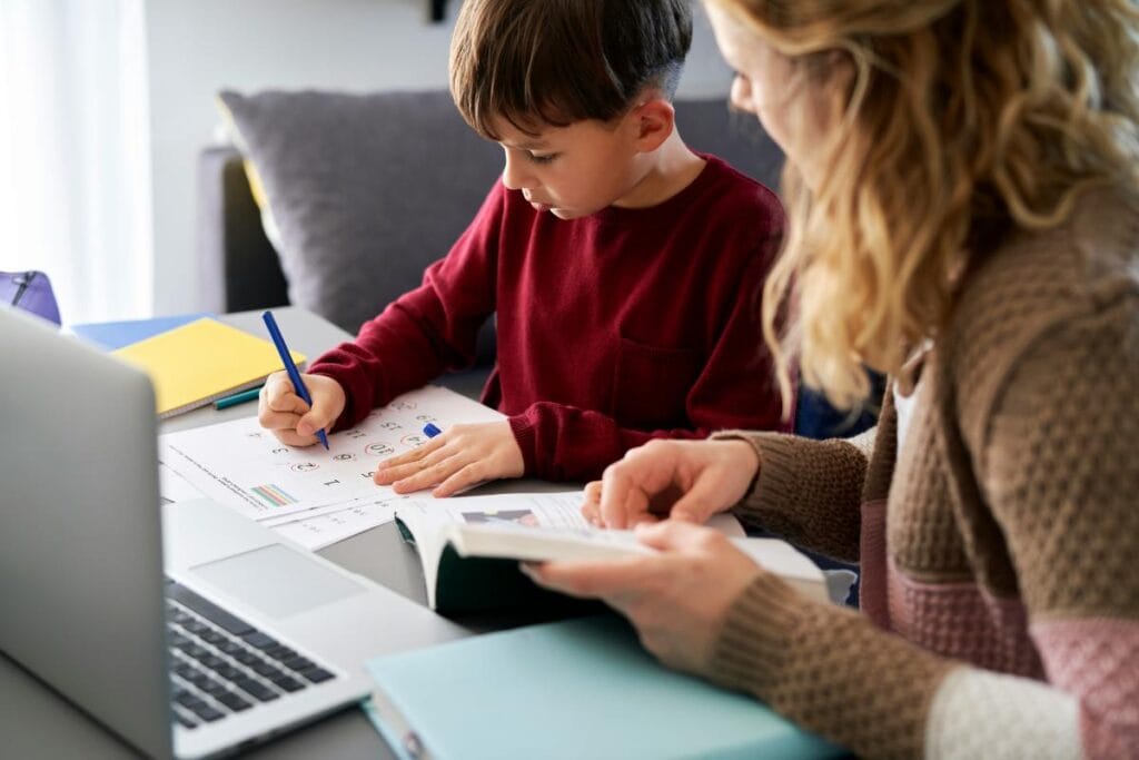 Parent helping a young child with homework at a desk, surrounded by books and a laptop, promoting productive study habits at home.