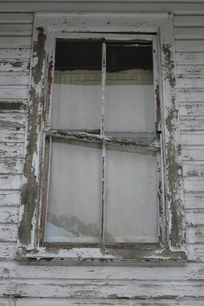 Weathered exterior window with peeling paint, warped wood, and advanced signs of rot on the frame and siding.