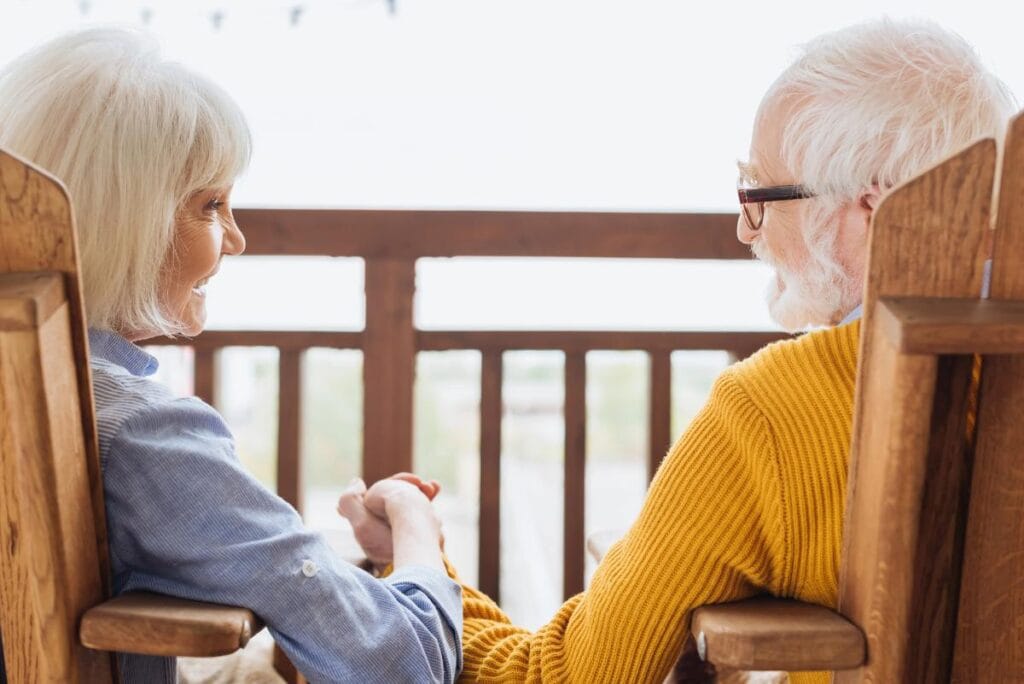 Senior couple sitting on porch rocking chairs holding hands, enjoying retirement security and peace of mind from reverse mortgage benefits.
