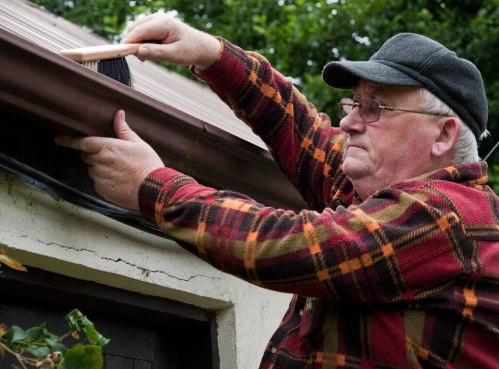 Senior homeowner cleaning roof gutters with a brush, performing seasonal roof maintenance to prevent water damage.