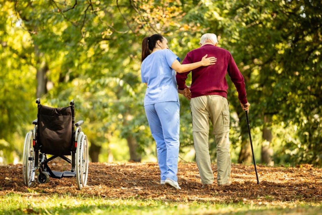 Care worker walking beside an elderly man with a cane in a wooded park, with an empty wheelchair nearby.