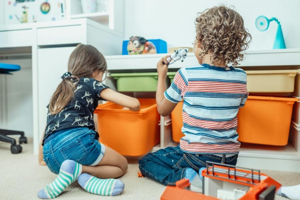 a boy and  a girl sitting in the floor pulling toys out of orange storage bins