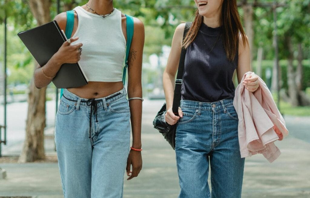Close-up of two students walking side by side outdoors, carrying books and bags, chatting and smiling on their way across campus.