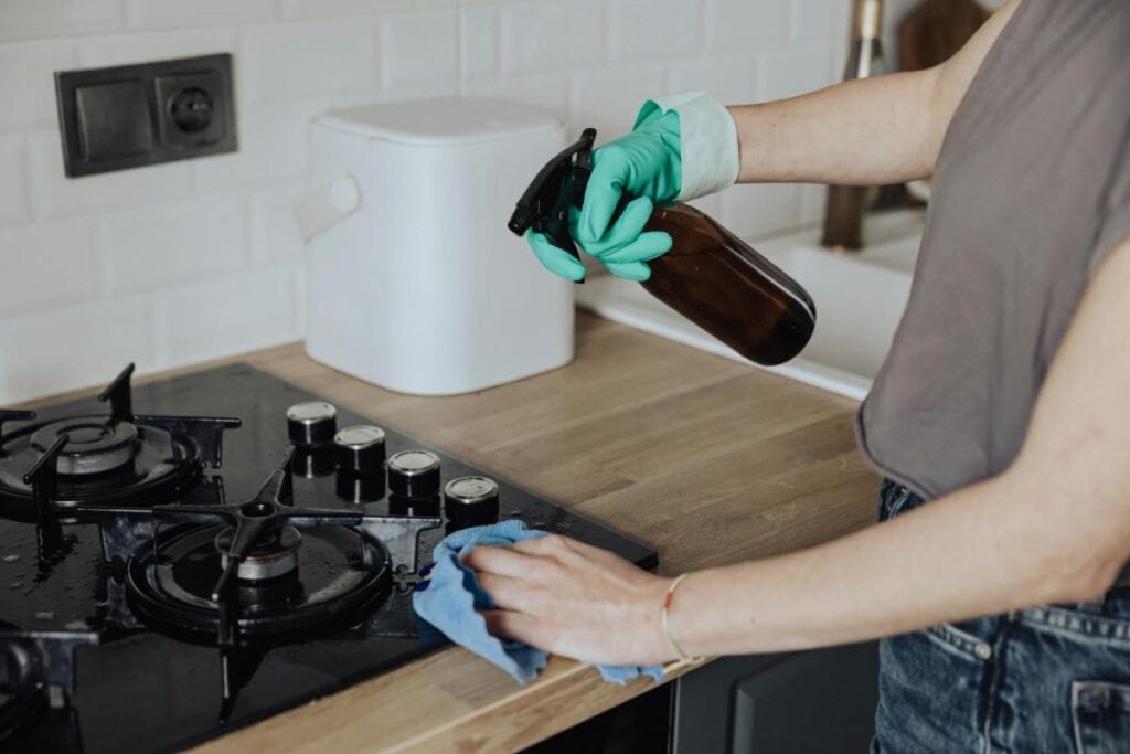 Person wearing green gloves sprays cleaner and wipes down a stovetop, focusing on grease removal from burners and surrounding surfaces.