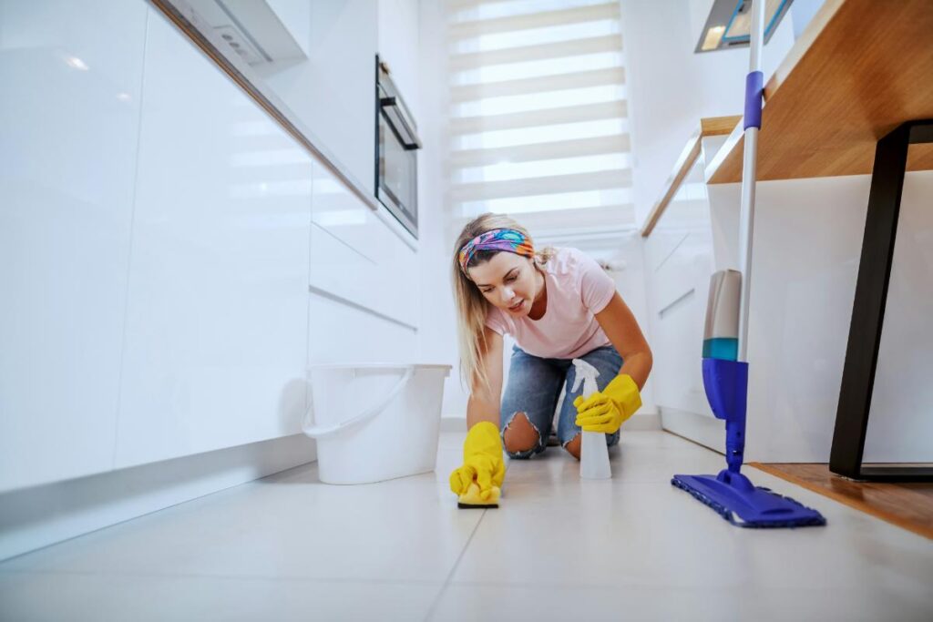 Woman wearing yellow gloves scrubs a kitchen floor with a sponge, surrounded by cleaning supplies, including a spray bottle, mop, and bucket.